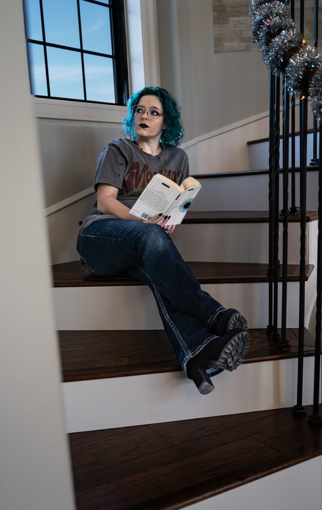 portrait of a woman with blue hair wearing glasses and dark lipstick, a grey shirt, dark pants, and black boots sitting on brown and white stairs holding a white and blue book staring off into the distance with a window behind her. Lyssa Benavidez Photography Elkhorn, NE Lyssab.com Self-portrait photography