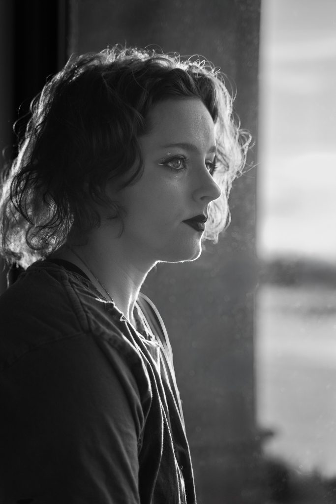 Black and white portrait of a woman wearing glasses and dark lipstick, a grey shirt standing in front of a window with a body of water in the background staring off into the distance. Lyssa Benavidez Photography Elkhorn, NE Lyssab.com Self-portrait photography