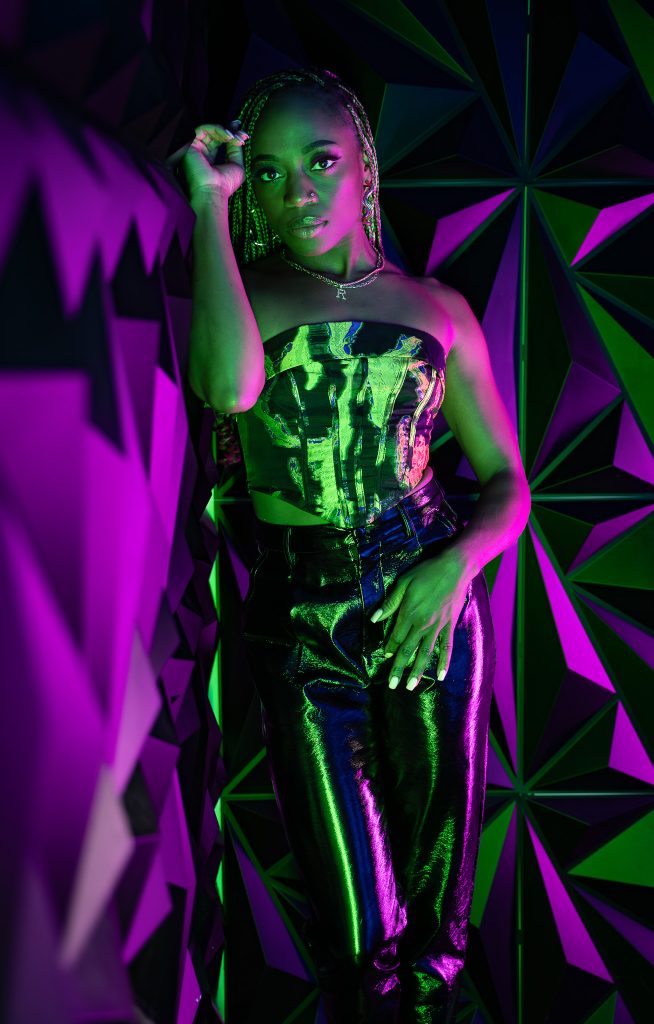 Creative light photo of a woman leaning against a wall wearing dark colored, shiny clothing with pink and green light and a geometric green and purple background. Lyssa Benavidez Photography Elkhorn, NE Lyssab.com studio photography. 