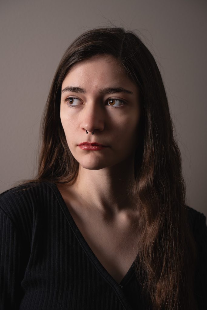 Dramatic portrait of a woman with long hair wearing a black shirt looking off into the distance standing in front of a grey background with dramatic lighting. Lyssa Benavidez Photography Elkhorn, NE Lyssab.com studio fine art photography