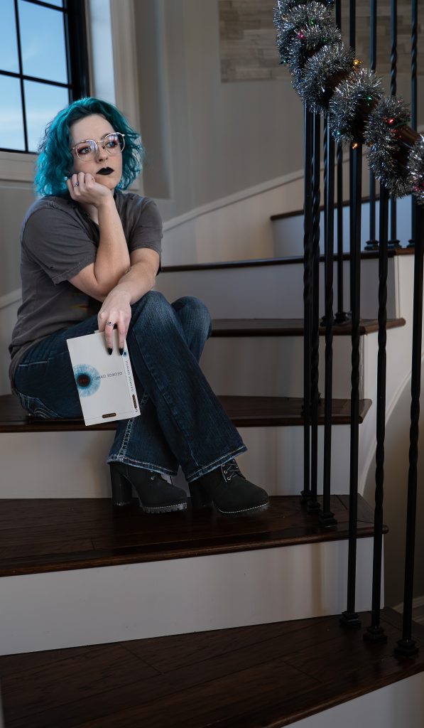 portrait of a woman with blue hair wearing glasses and dark lipstick, a grey shirt, dark pants, and black boots sitting on brown and white stairs holding a white and blue book staring off into the distance with a window behind her. Lyssa Benavidez Photography Elkhorn, NE Lyssab.com Self-portrait photography