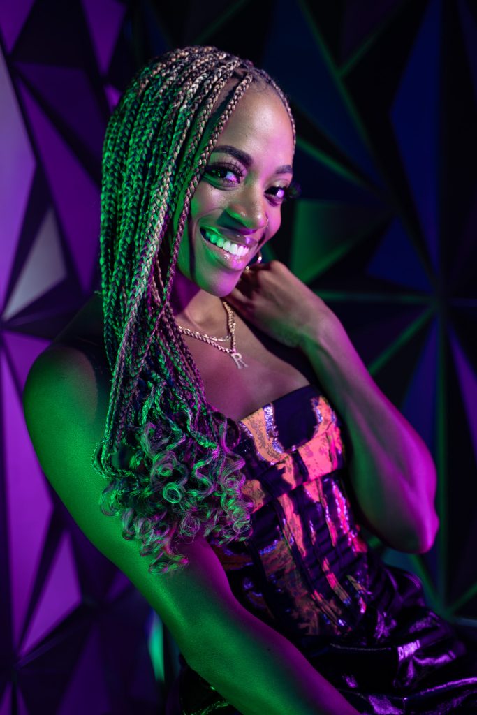 Creative light photo of a woman sitting on a stool wearing dark colored, shiny clothing with pink and green light and a geometric green and purple background. Lyssa Benavidez Photography Elkhorn, NE Lyssab.com studio photography. 