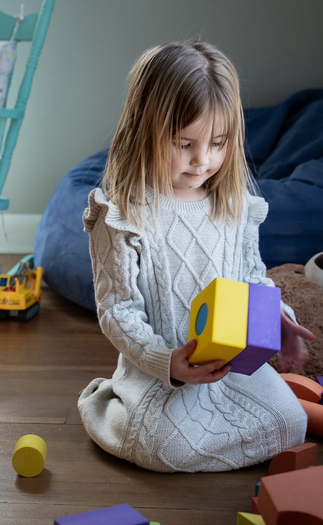 Lifestyle portrait of a little girl with blonde hair playing with bright colored toys wearing a grey dress sitting on the floor surrounded by toys and chairs. Lyssa Benavidez Photography Elkhorn, NE Lyssab.com lifestyle family photography. 