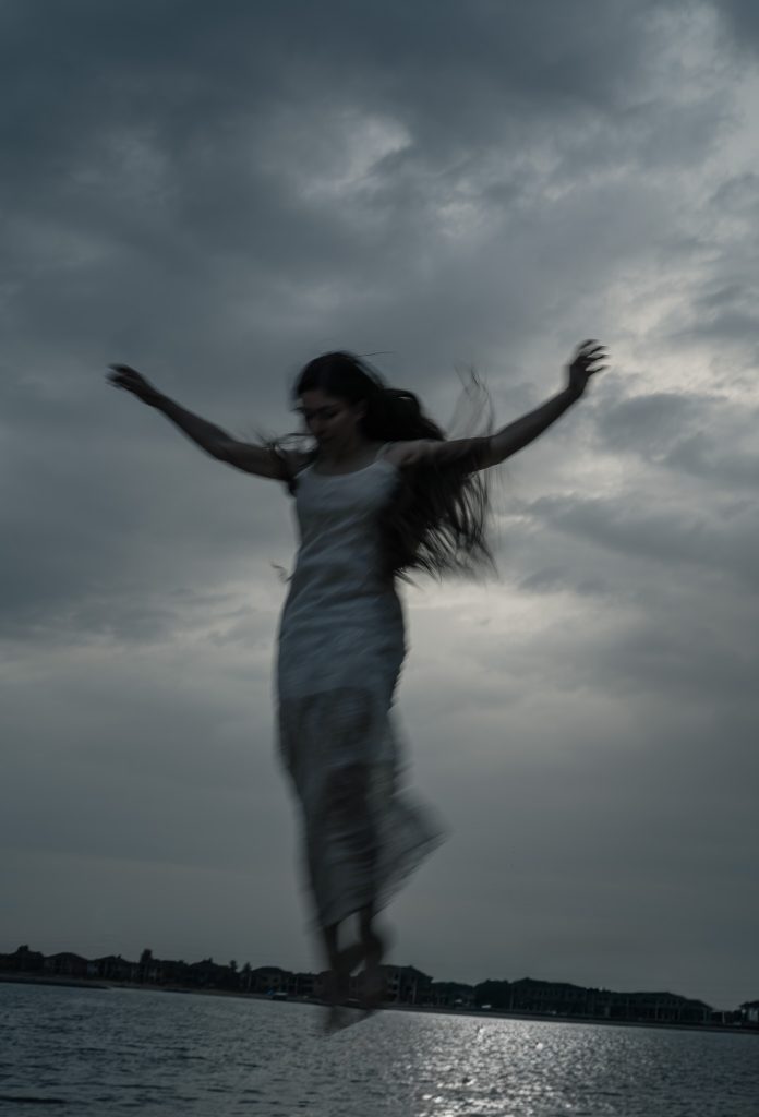 Surrealist portrait of a woman who appears to be flying with her arms stretched out, looking down with a dark blue water and cloudy sky behind her.  Lyssa Benavidez Photography Elkhorn, NE Lyssab.com Fine Art Photography Surrealist photography