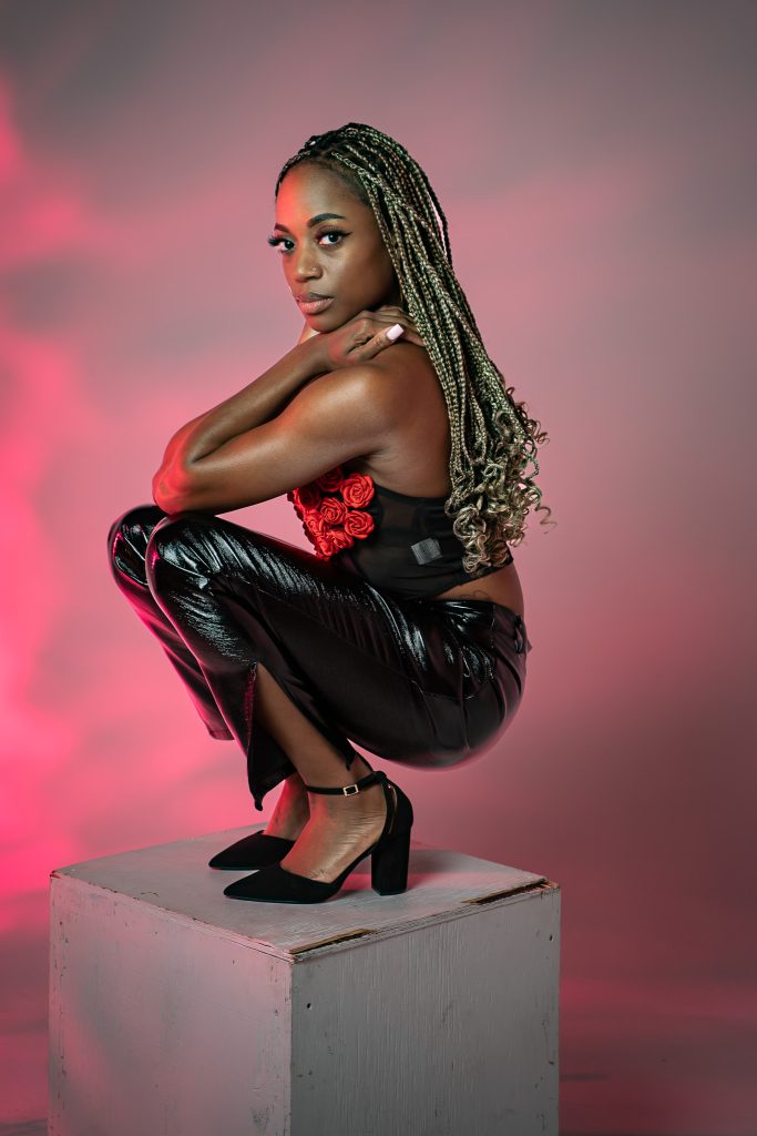 Creative light photo of a woman sitting on a white box wearing black and red colored, shiny clothing with a red background. Lyssa Benavidez Photography Elkhorn, NE Lyssab.com studio photography. 