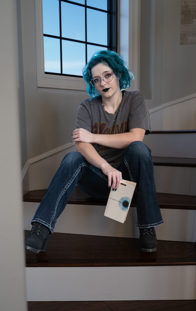 portrait of a woman with blue hair wearing glasses and dark lipstick, a grey shirt, dark pants, and black boots sitting on brown and white stairs holding a white and blue book staring into the camera with a window behind her. Lyssa Benavidez Photography Elkhorn, NE Lyssab.com Self-portrait photography