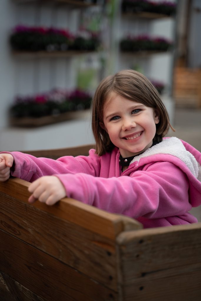 Portrait of a little girl smiling and wearing a pink shirt, sitting backwards in a sleigh with plants in the background. Lyssa Benavidez Photography Elkhorn, NE Lyssab.com Lifestyle family photography