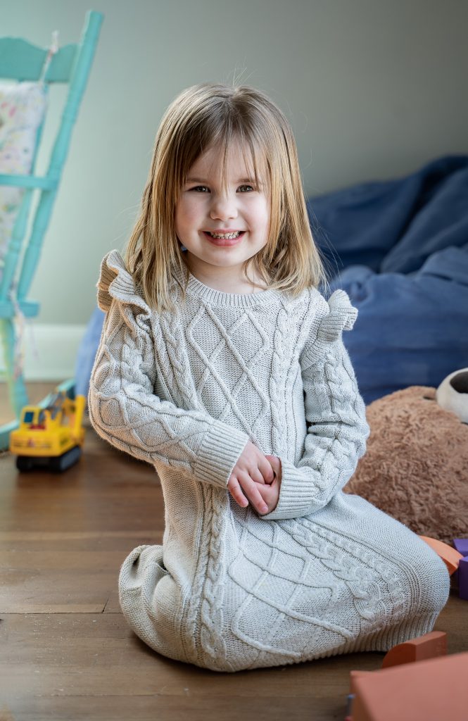 Lifestyle portrait of a little girl with blonde hair smiling wearing a grey dress sitting on the floor surrounded by toys and chairs. Lyssa Benavidez Photography Elkhorn, NE Lyssab.com lifestyle family photography. 