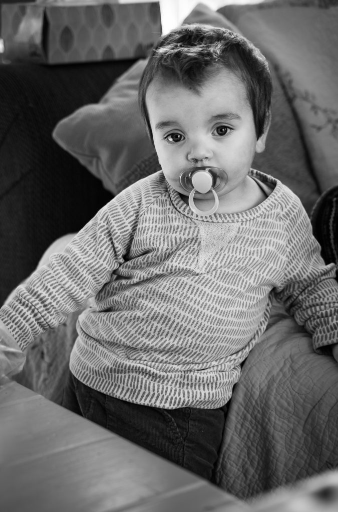Black and white photo of a baby boy wearing a white and grey striped shirt and dark pants standing between a table and a couch with pillows on it. Lyssa Benavidez Photography Elkhorn, NE Lyssab.com lifestyle family photography 