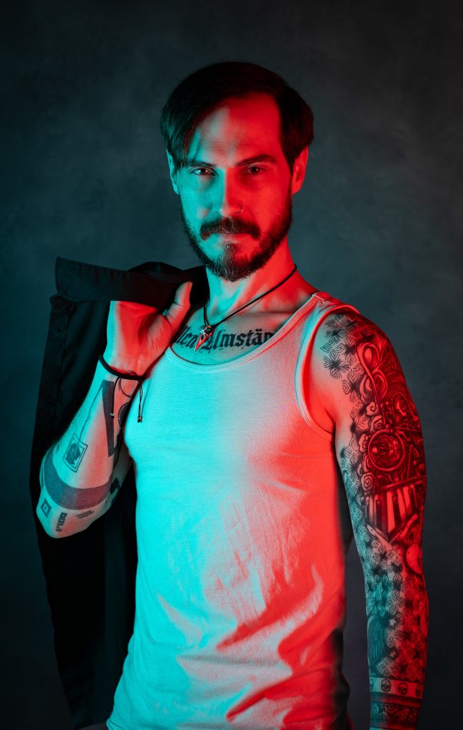 Creative portrait of a man with a menacing look wearing a white tank top, holding a black shirt with red and blue creative lighting standing in front of a dark blue background. Lyssa Benavidez Photography Elkhorn, NE studio photography. 