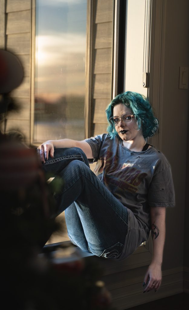 portrait of a woman with blue hair wearing glasses and dark lipstick, a grey shirt, dark pants, and black boots sitting in a windowsill with a window behind her. Lyssa Benavidez Photography Elkhorn, NE Lyssab.com Self-portrait photography