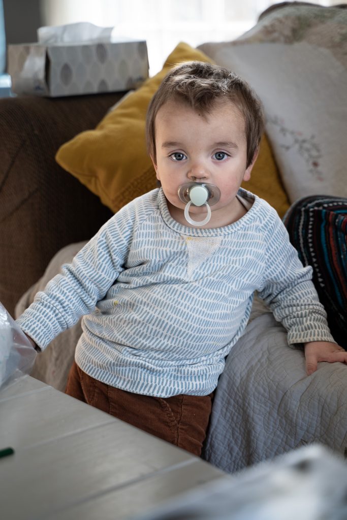 Lifestyle portrait of a baby boy wearing a white and blue striped shirt and brown pants standing between a table and a couch with pillows on it. Lyssa Benavidez Photography Elkhorn, NE Lyssab.com lifestyle family photography 