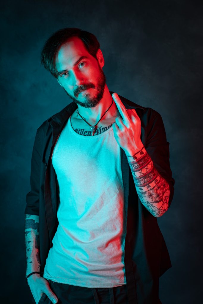 Creative portrait of a man flipping off the camera wearing a black and white shirt with his other hand in his pocket, with red and blue creative lighting standing in front of a dark blue background. Lyssa Benavidez Photography Elkhorn, NE studio photography. 