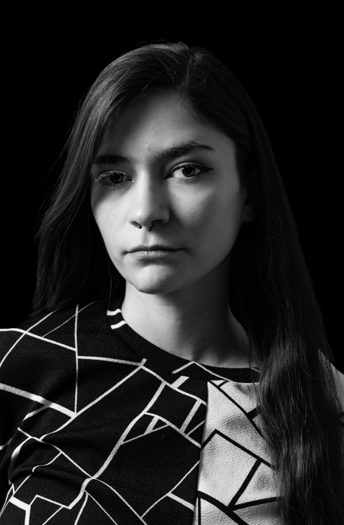 Black & white portrait of a woman with long hair wearing a black and white shirt looking at the camera standing in front of a black background with dramatic lighting. Lyssa Benavidez Photography Elkhorn, NE Lyssab.com studio fine art photography