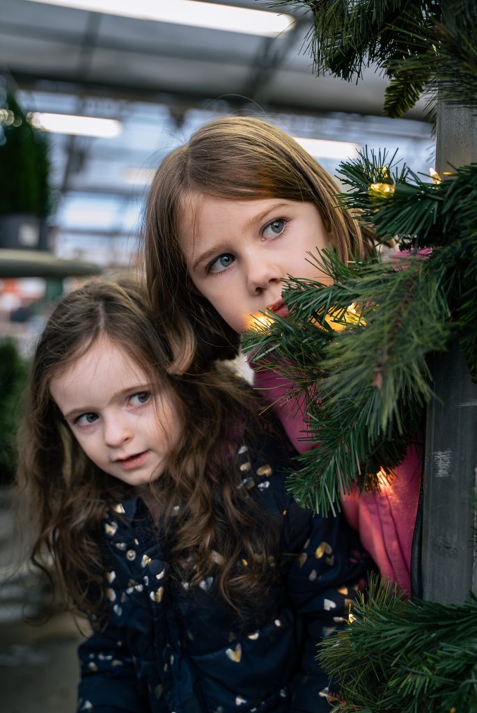 Portrait of two girls peeking around a Christmas tree together with plants in the background. Lyssa Benavidez Photography Elkhorn, NE Lyssab.com Lifestyle family photography