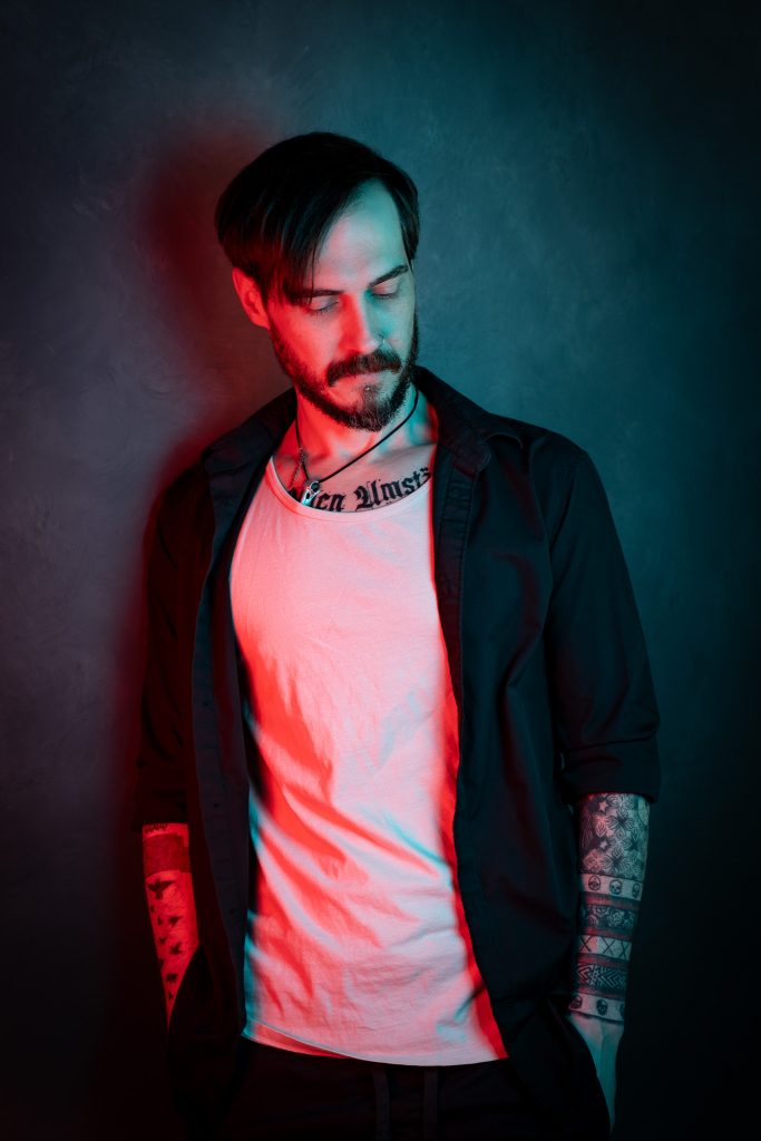 Creative portrait of a man looking down wearing a black and white shirt, with red and blue creative lighting standing in front of a dark blue background. Lyssa Benavidez Photography Elkhorn, NE studio photography. 