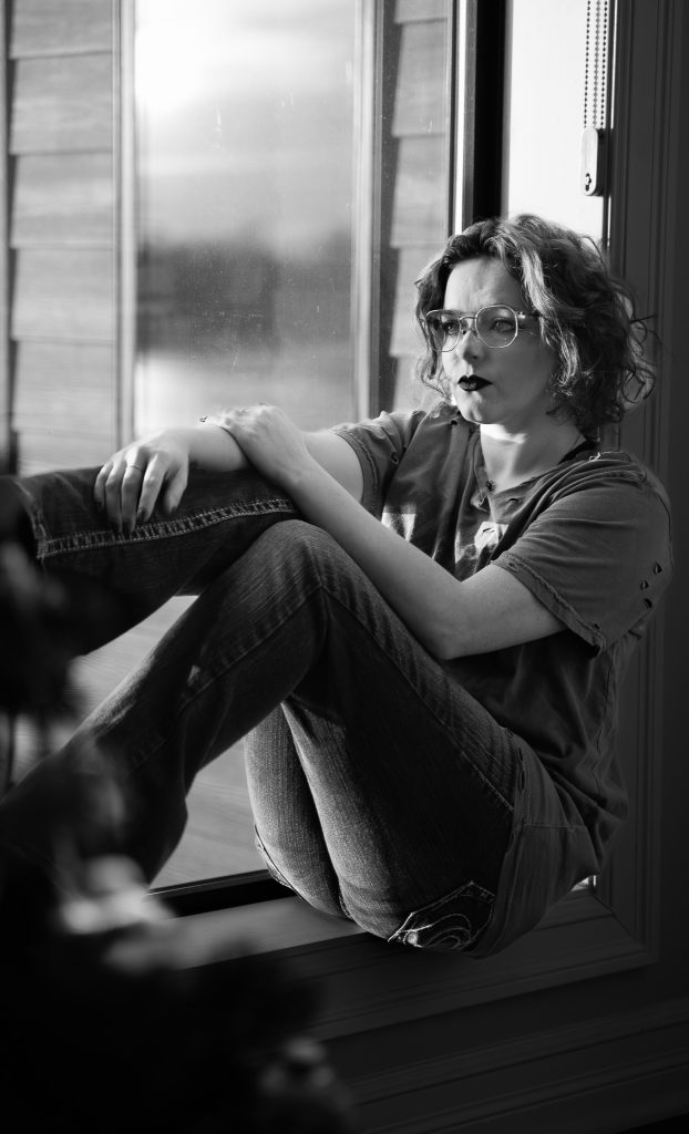 Black and white portrait of a woman wearing glasses and dark lipstick, a grey shirt, dark pants, and black boots sitting in a windowsill. Lyssa Benavidez Photography Elkhorn, NE Lyssab.com Self-portrait photography