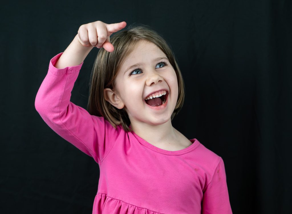 Studio portrait of a little girl with brown hair who is pointing to something off camera with a big smile on her face wearing a pink dress standing in front of a black background. Lyssa Benavidez Photography Elkhorn NE Lyssab.com studio photography. 