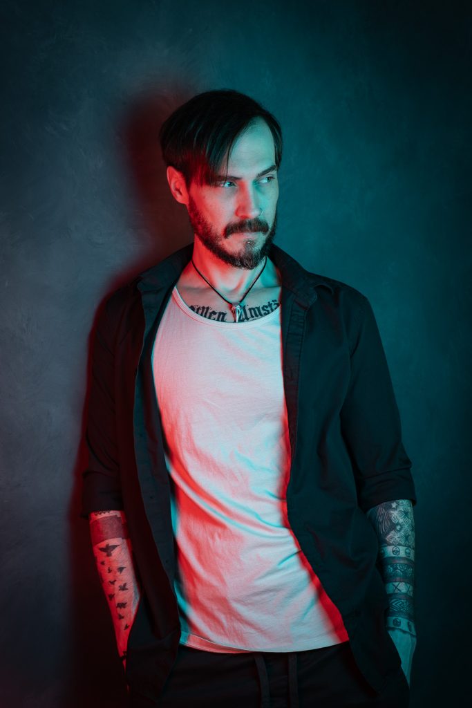 Creative light portrait of a man wearing a black and white shirt, with red and blue creative lighting standing in front of a dark blue background. Lyssa Benavidez Photography Elkhorn, NE studio photography. 