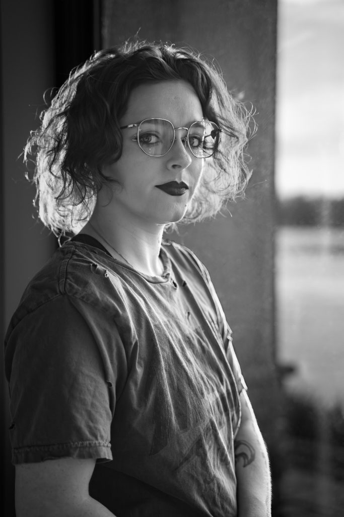 Black and white portrait of a woman wearing glasses and dark lipstick, a grey shirt standing in front of a window with a body of water in the background looking at the camera. Lyssa Benavidez Photography Elkhorn, NE Lyssab.com Self-portrait photography