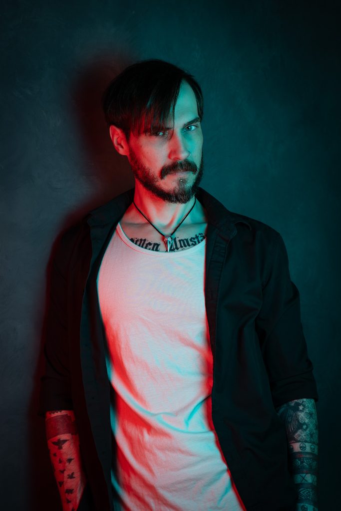 Creative portrait of a man with a menacing look wearing a black and white shirt, with red and blue creative lighting standing in front of a dark blue background. Lyssa Benavidez Photography Elkhorn, NE studio photography. 