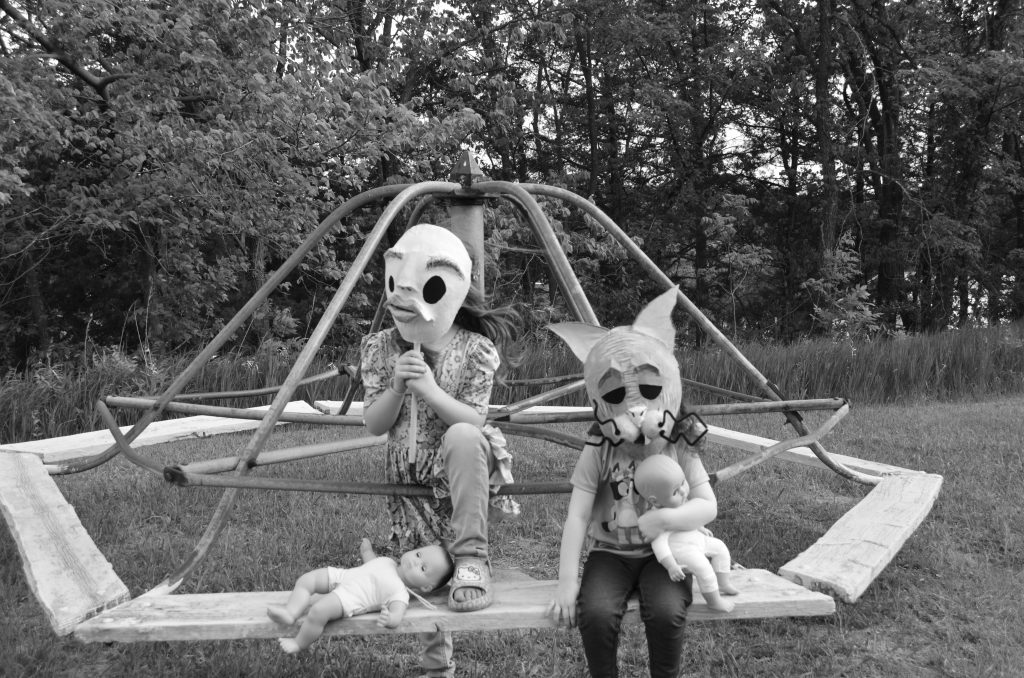 Black and white photo of two kids wearing creepy masks playing with baby dolls sitting on an old merry-go-round with trees in the background